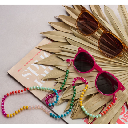 Bright pink sunglasses with dark lenses and a colorful beaded strap, styled on a book with dried palm leaves and another pair of sunglasses on a light gray background.