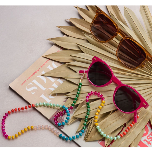 Bright pink sunglasses with dark lenses and a colorful beaded strap, styled on a book with dried palm leaves and another pair of sunglasses on a light gray background.