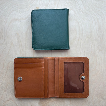 Two leather bifold wallets shown on a light wood background: one closed in hunter green at the top and one open in tan brown at the bottom, displaying card slots and an ID window.