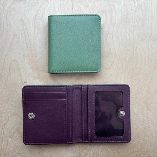 Two leather bifold wallets shown on a light wood background: one closed in sage green at the top and one open in deep purple at the bottom, displaying card slots, a snap closure, and an ID window.