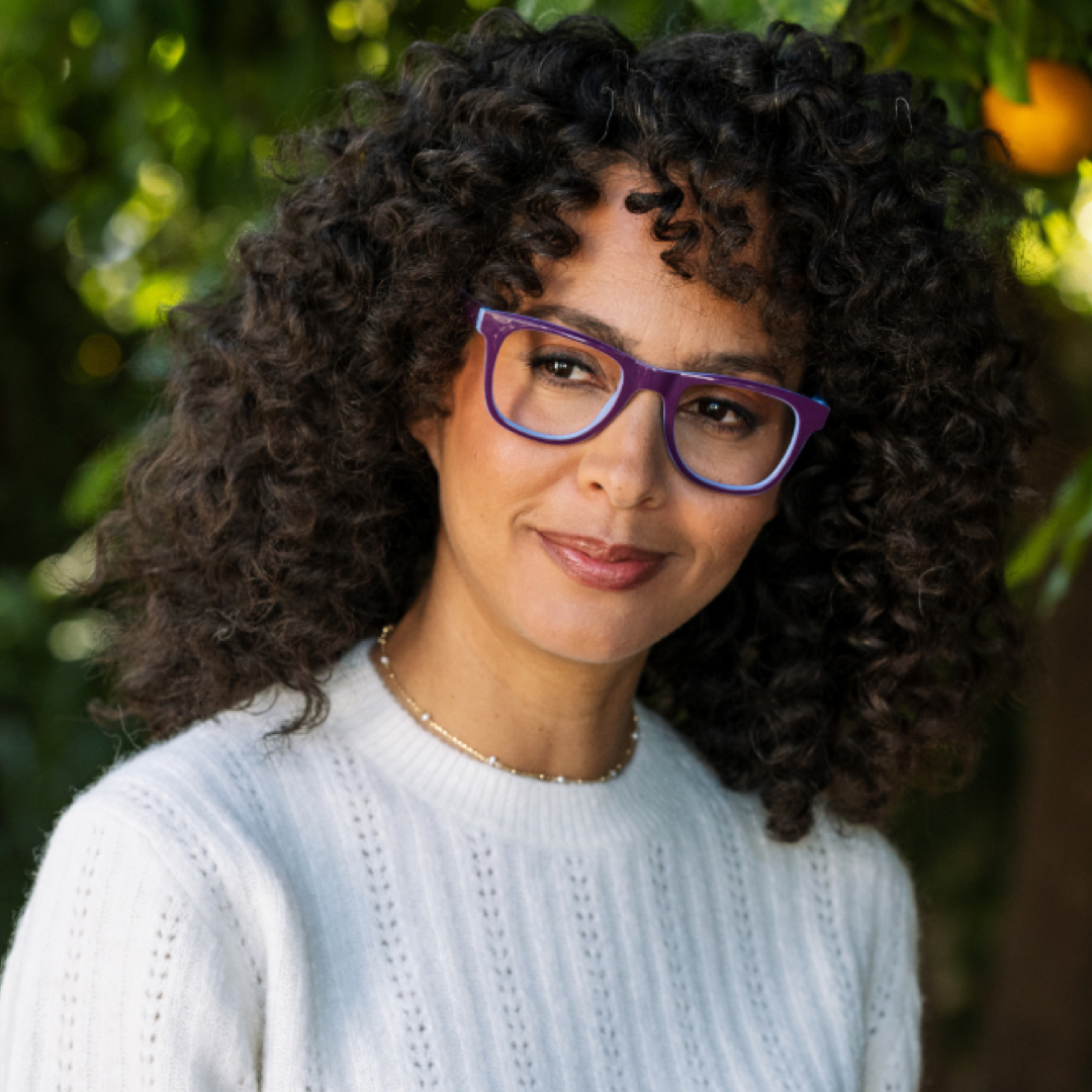 A close-up portrait of a woman with curly dark hair wearing purple-framed eyeglasses and a white knit sweater, photographed outdoors with blurred green foliage and an orange in the background.
