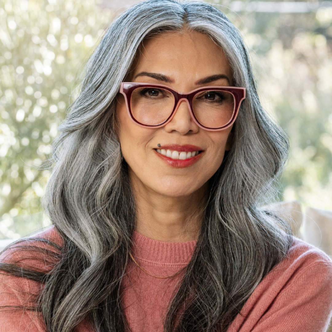A close-up portrait of a woman with long gray hair wearing cat-eye glasses with burgundy frames and a dusty rose sweater, set against a softly blurred outdoor background.