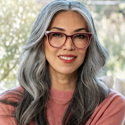 A close-up portrait of a woman with long gray hair wearing cat-eye glasses with burgundy frames and a dusty rose sweater, set against a softly blurred outdoor background.