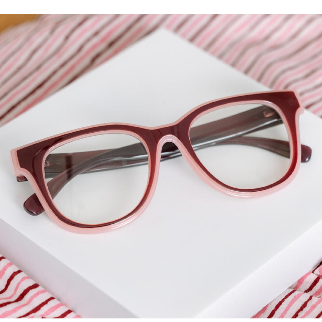 A pair of chunky, oversized cat-eye eyeglasses with a translucent dusty rose and deep burgundy frame, resting on a white rectangular box with pink and white striped fabric in the background.