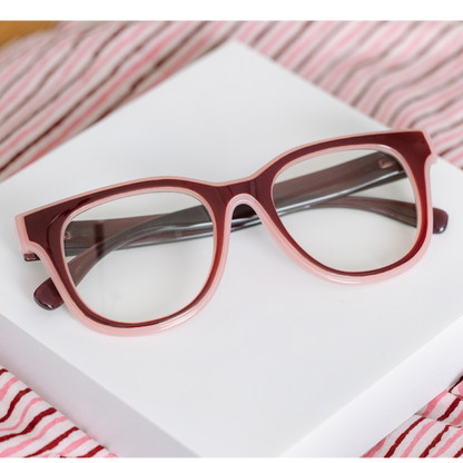 A pair of chunky, oversized cat-eye eyeglasses with a translucent dusty rose and deep burgundy frame, resting on a white rectangular box with pink and white striped fabric in the background.