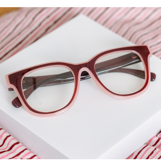 A pair of chunky, oversized cat-eye eyeglasses with a translucent dusty rose and deep burgundy frame, resting on a white rectangular box with pink and white striped fabric in the background.