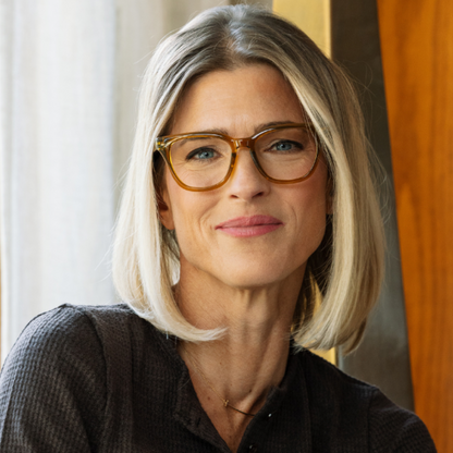 A close-up portrait of a woman with shoulder-length blonde hair wearing tortoiseshell eyeglasses and a dark textured top, photographed indoors with soft natural light.