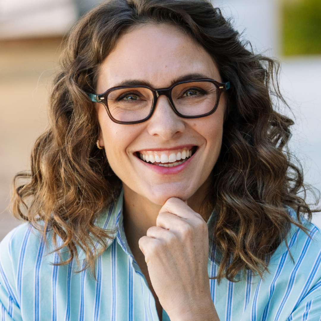 A smiling woman with wavy brown hair and black-rimmed eyeglasses, wearing a light blue and white striped button-up shirt, photographed in a bright outdoor setting.