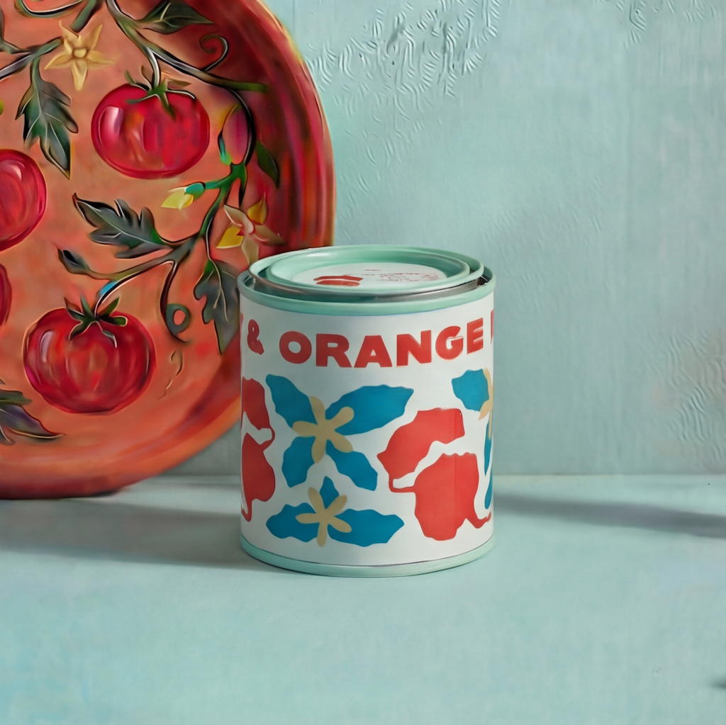 Candle in a tin vessel labeled 'Beet & Orange'  next to a plate with tomato pattern on a light blue background