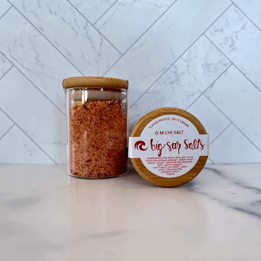 Glass container of O M Chi Big Sur Salts on marble counter, against gray tile background