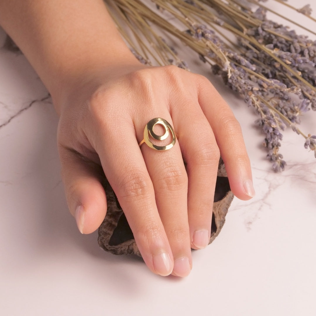 Hand wearing a brass ring with an open circle swirl with lavender flowers in the background