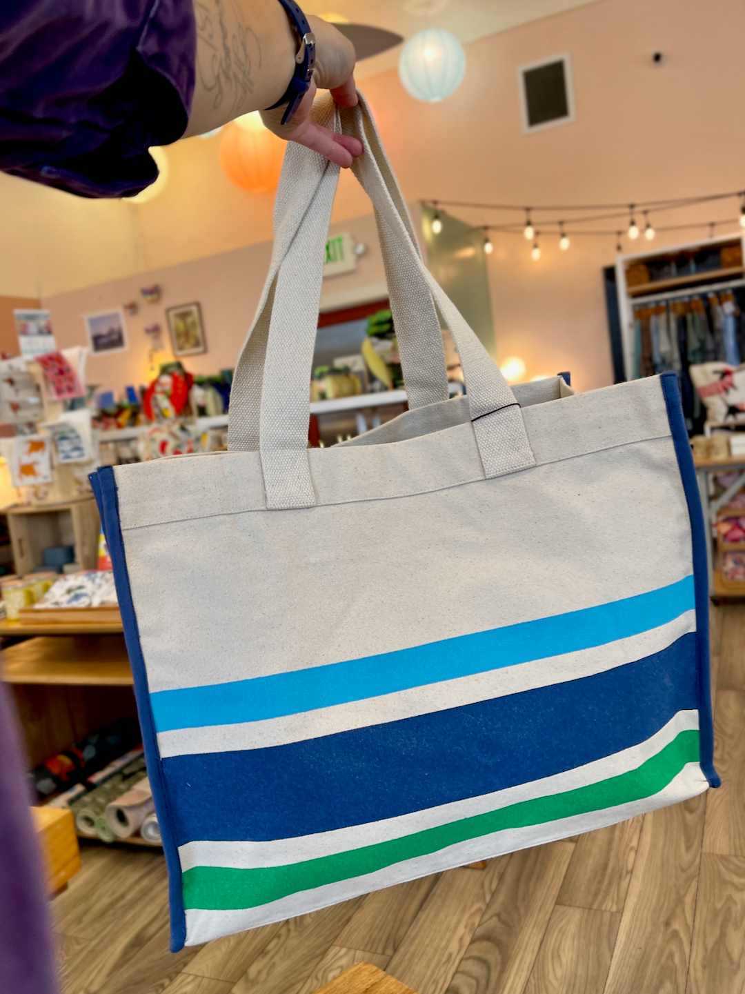 Arm holding out blue and green striped tote. Shop interior in background.