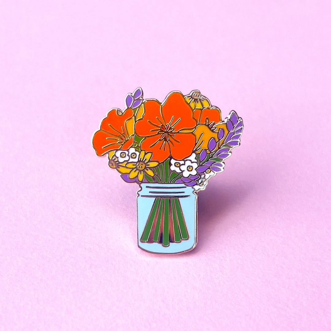 Floral design pin with orange poppies, lavender sprigs, and yellow flowers in a blue jar on a pink background