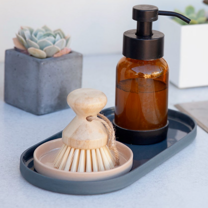 Brown soap dispenser and wooden brush on a denim colored tray with a succulent plant in the background.