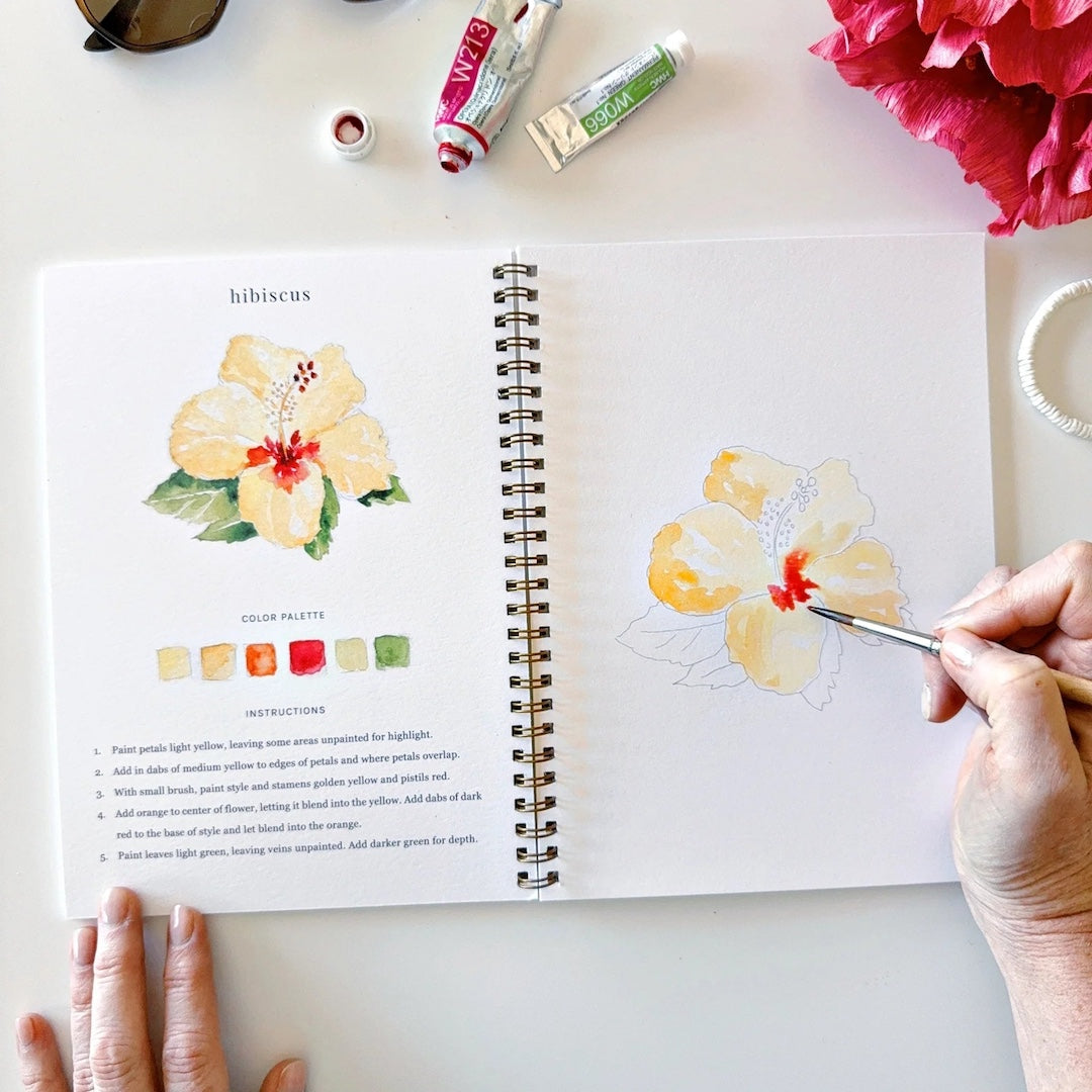Person painting a hibiscus flower in a sketchbook with art supplies on a table.
