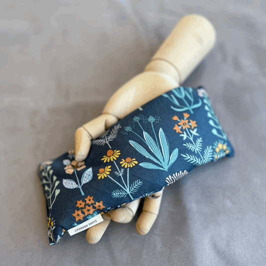 a wooden mannequin hand holding a rectangular eye pillow that has a floral pattern on a navy background