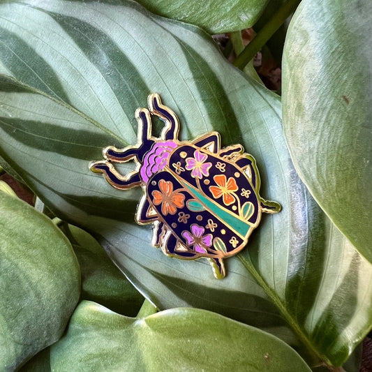 beetle shaped enamel pin with a floral pattern on it pinned to a green leaf