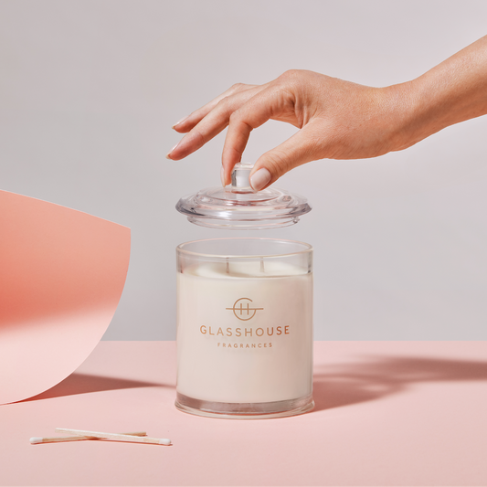 Glasshouse Fragrances candle with a hand reaching to touch the lid on a pink and gray background