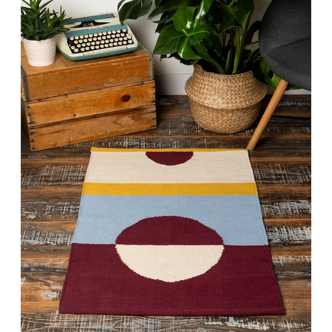 Colorful geometric rug on a wooden floor with plants and a typewriter in the background
