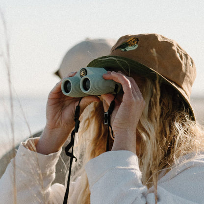 Person wearing a brown hat and white jacket using green binoculars in a natural setting.