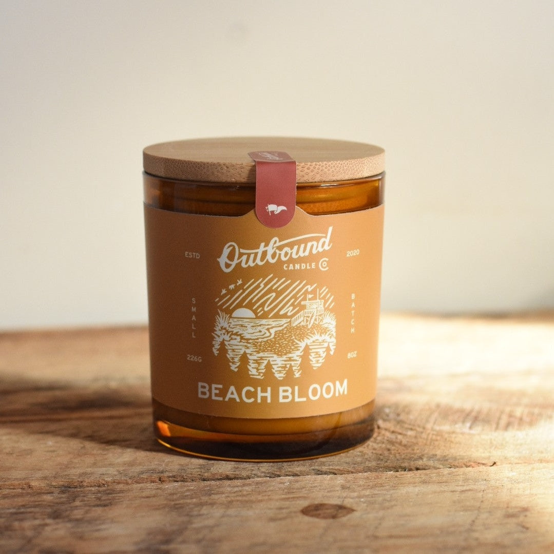 Candle labeled 'Beach Bloom' by 'Outbound Candle Co.' on a wooden surface.