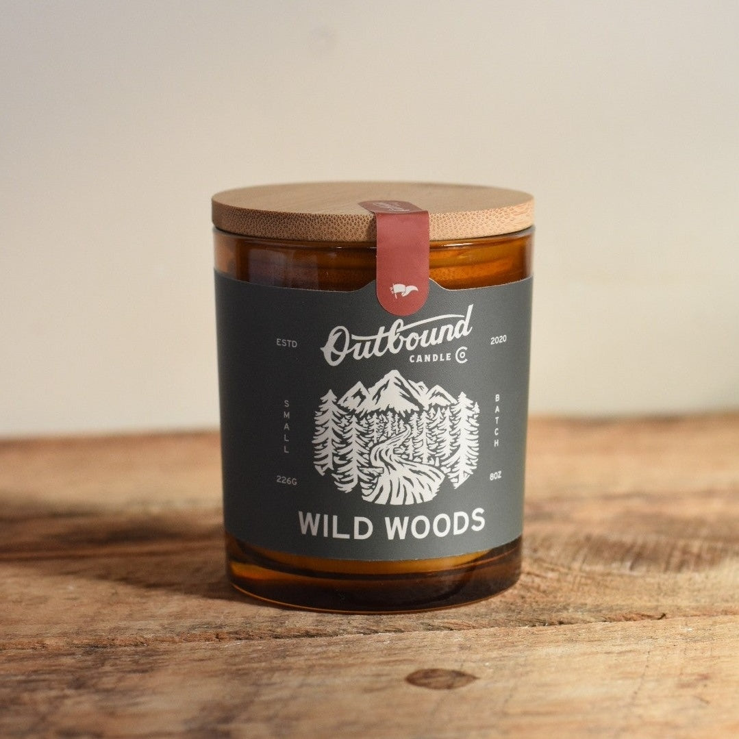 Candle jar labeled 'Wild Woods' by 'Outbound Candle Co.' on a wooden surface.