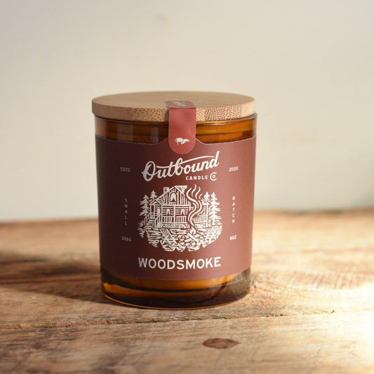 Candle jar labeled 'Outbound Candle Co. Woods smoke' on a wooden surface