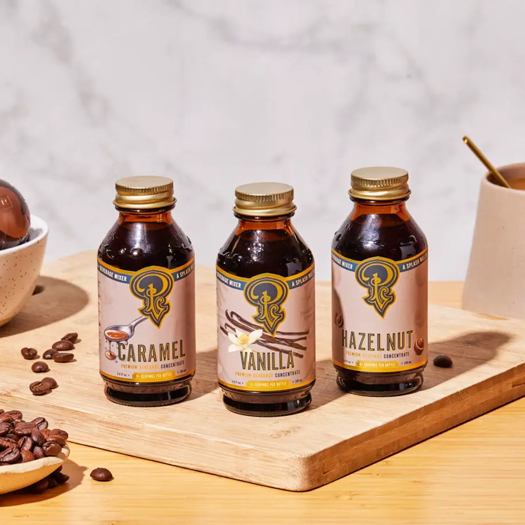 Three bottles of caramel, vanilla, and hazelnut flavor concentrates on a wooden board with coffee beans and a cup.