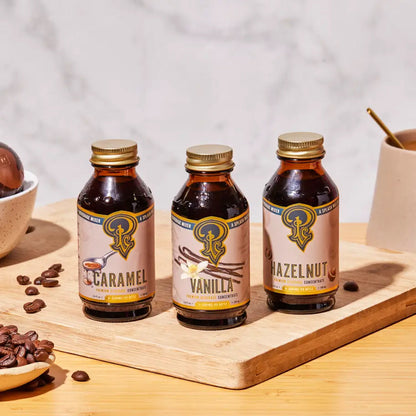Three bottles of caramel, vanilla, and hazelnut flavor concentrates on a wooden board with coffee beans and a cup.