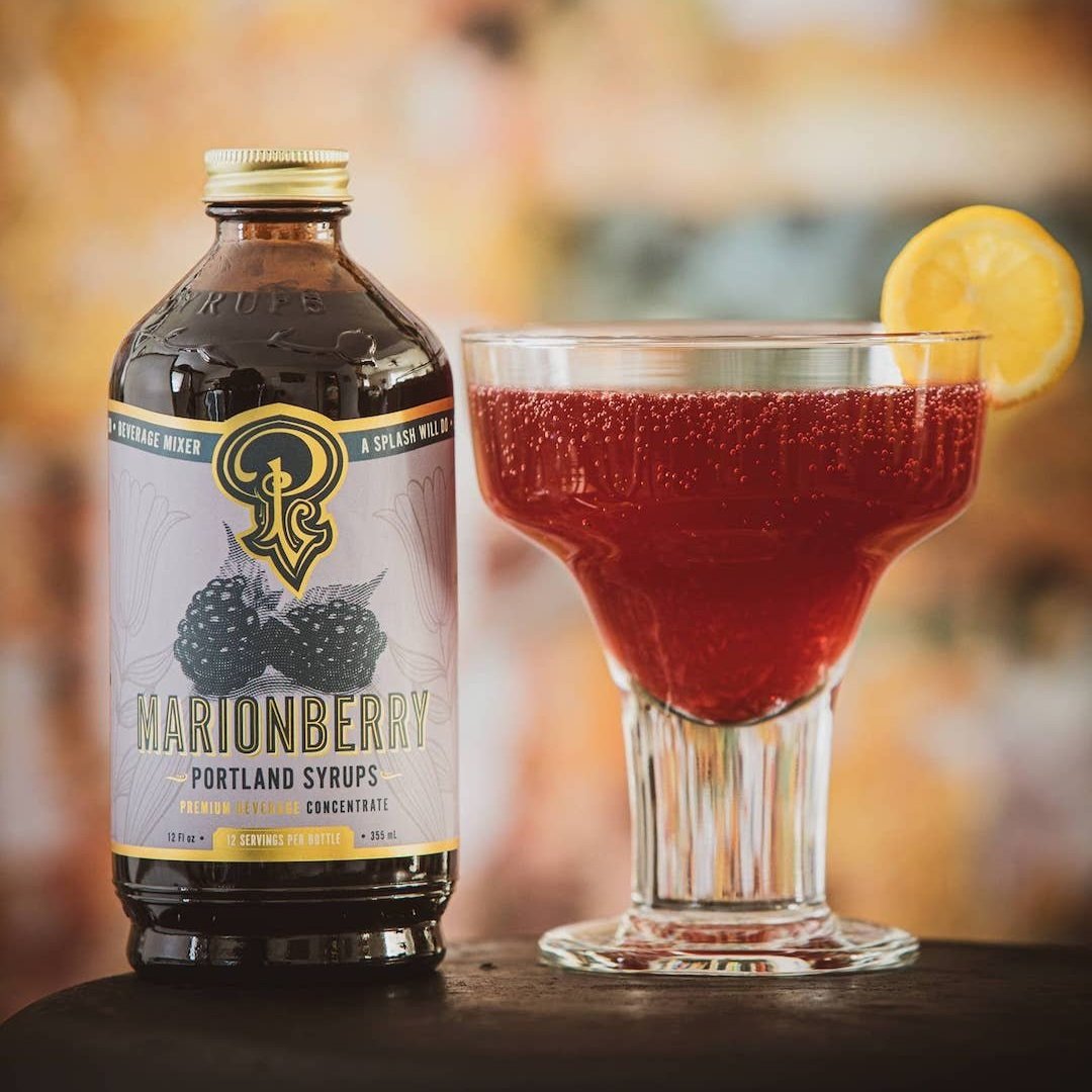 Marionberry syrup bottle next to a cocktail with a lemon twist on a blurred bar background