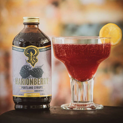 Marionberry syrup bottle next to a cocktail with a lemon twist on a blurred bar background