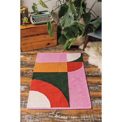 Colorful geometric rug on a wooden floor with plants and a typewriter in the background