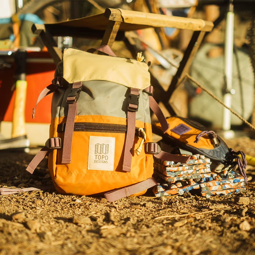 Orange and beige backpack with 'Topo Designs' logo on a dirt ground.