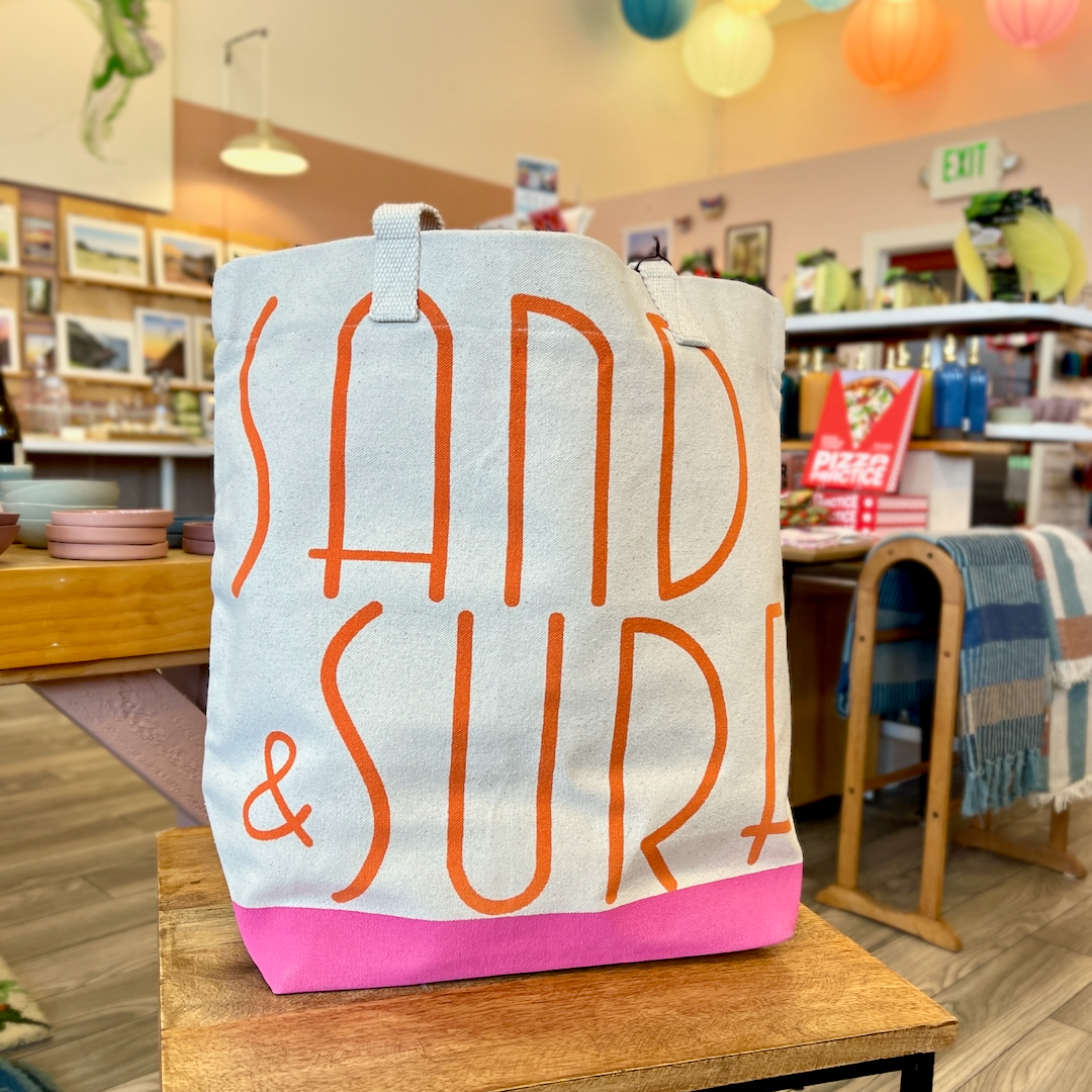 Canvas tote with orange lettering that spells out Sand & Surf. Tote has pink bottom. Tote is sitting on a wooden table, with store interior in the background.