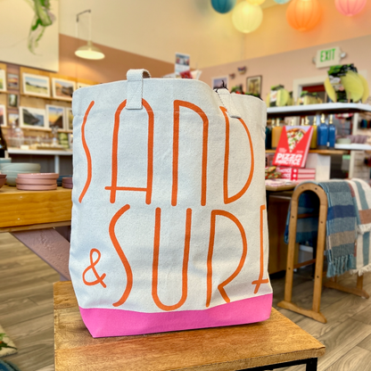 Canvas tote with orange lettering that spells out Sand & Surf. Tote has pink bottom. Tote is sitting on a wooden table, with store interior in the background.