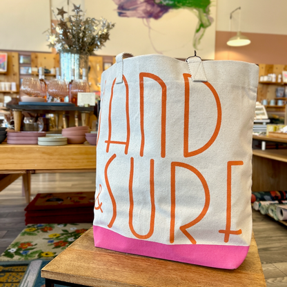 Side angle of canvas tote with orange lettering that spells out Sand & Surf. Tote has pink bottom. Tote is sitting on a wooden table, with store interior in the background.
