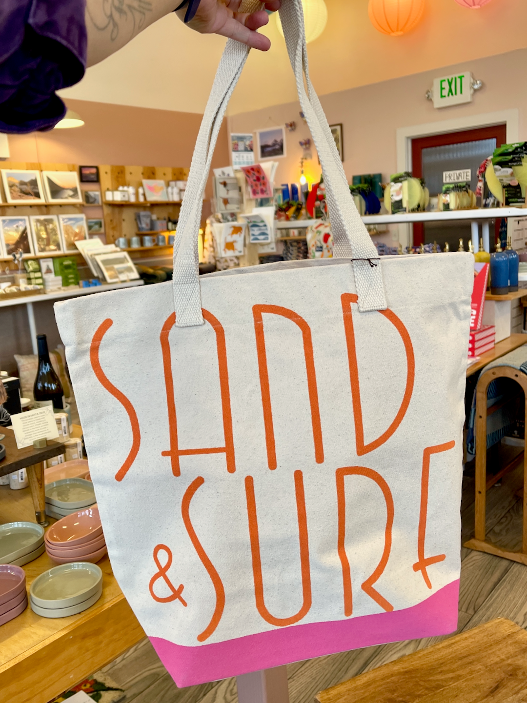 Canvas tote with orange lettering that spells out Sand & Surf. Tote has pink bottom. Tote is being held up by an arm. Store interior in background.