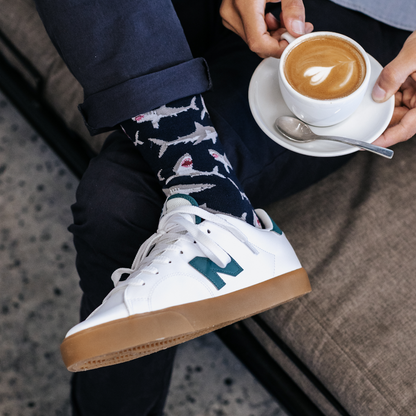 Person wearing shark-patterned socks with white sneakers, holding a cup of coffee.