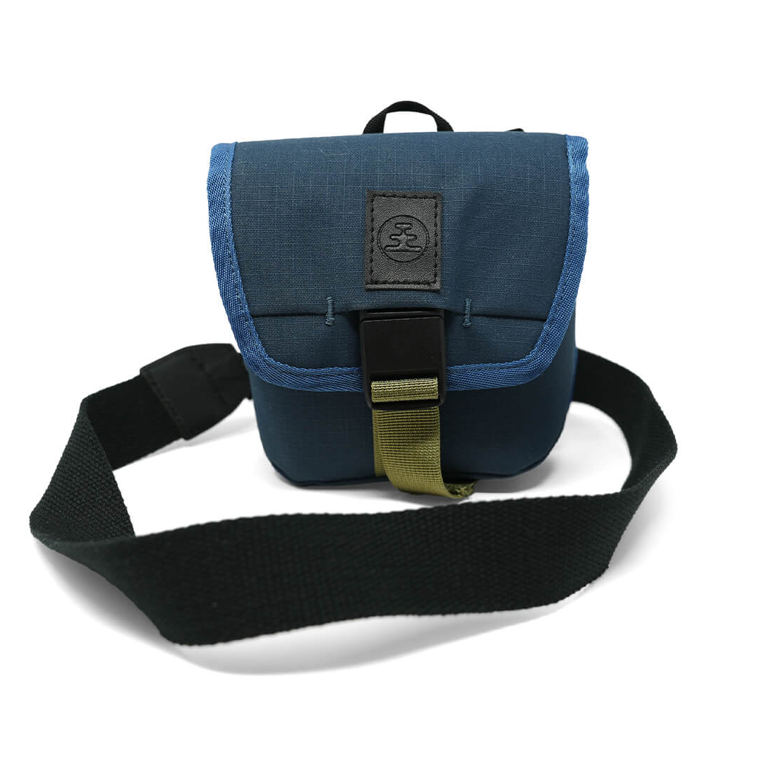 A blue fabric crossbody bag with a black strap, black plastic buckle, and a black leather patch on the front flap, photographed on a white background.