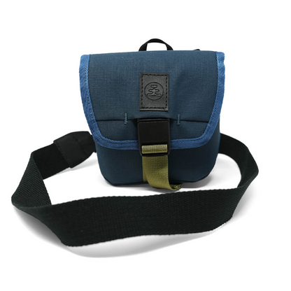 A blue fabric crossbody bag with a black strap, black plastic buckle, and a black leather patch on the front flap, photographed on a white background.