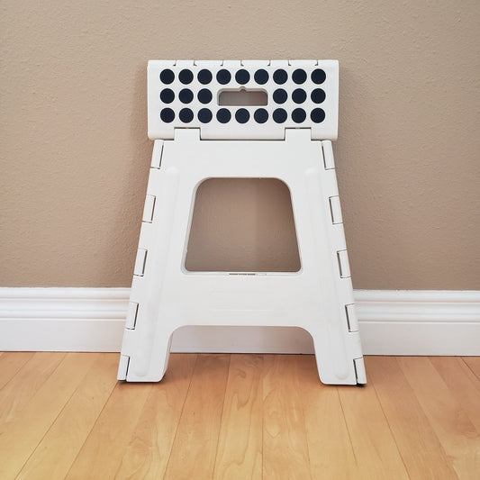 White folding stool with black dots on a wooden floor folded up against a beige wall.
