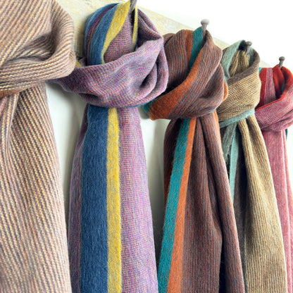 Several woven scarves in muted striped colors hang from hooks on a light wall. The visible scarves include beige, blue with yellow and purple stripes, brown with orange and teal stripes, taupe, and dusty pink.