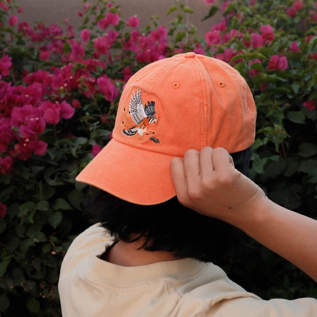 Person wearing an orange cap with a kestrel design, standing in front of pink flowers.