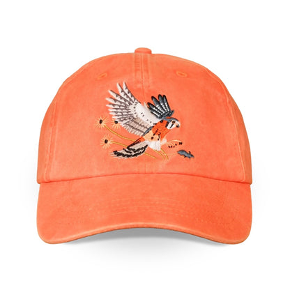An orange distressed baseball cap featuring an embroidered American kestrel with wings spread, swooping down to catch a mouse, on a clean white background.
