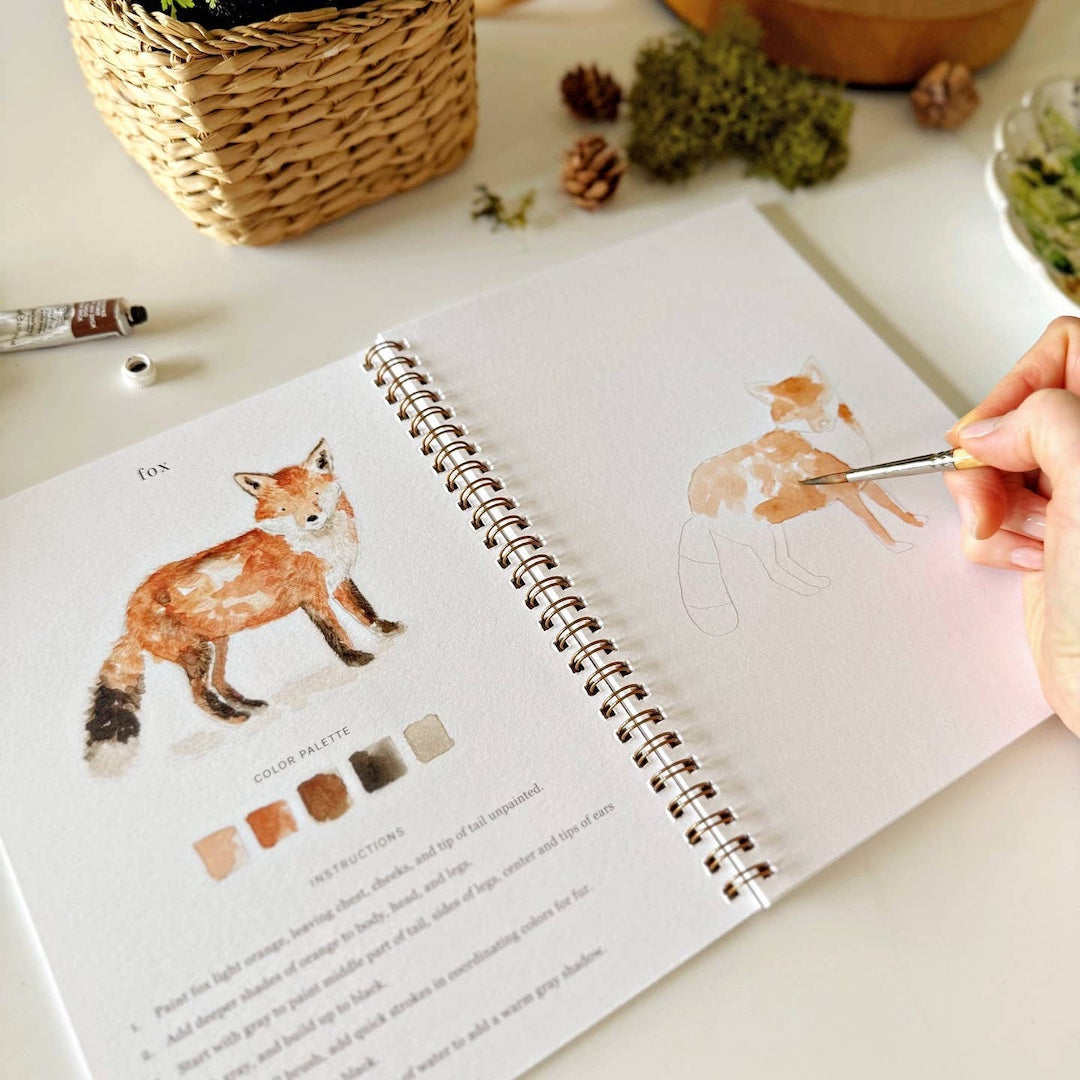 Person painting a fox illustration in a sketchbook with a paintbrush.