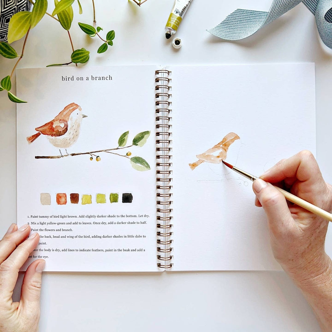 Person painting a bird illustration in a book with a paintbrush.