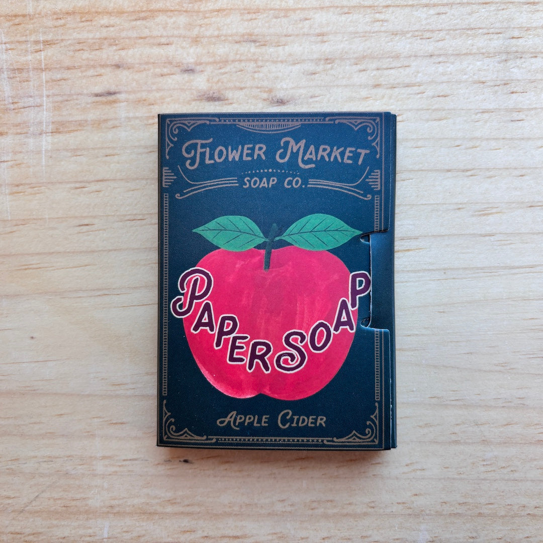 A vintage-style soap box for Flower Market Paper Soap, Apple Cider scent, resting on a light wood surface. The box is dark teal with a large red apple graphic and green leaves, featuring the brand name 'Flower Market Soap Co.
