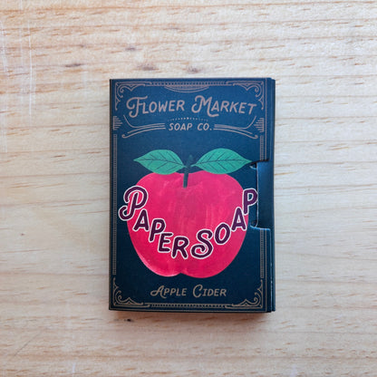 A vintage-style soap box for Flower Market Paper Soap, Apple Cider scent, resting on a light wood surface. The box is dark teal with a large red apple graphic and green leaves, featuring the brand name 'Flower Market Soap Co.