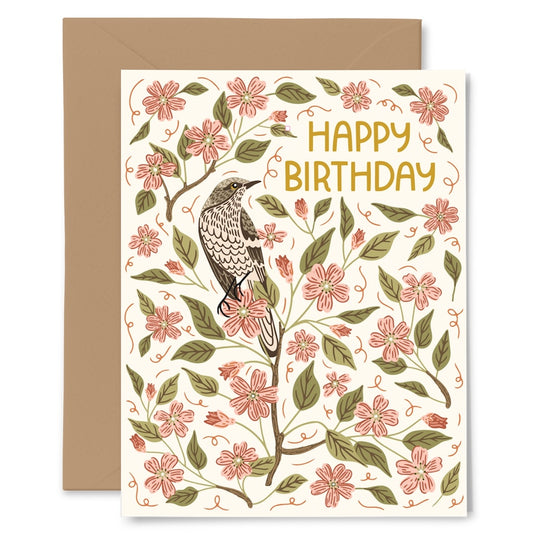 A floral and bird birthday card with the words “HAPPY BIRTHDAY” in gold lettering, shown with a brown envelope behind it on a white background.