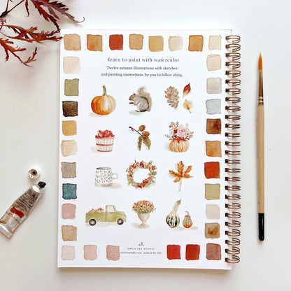Artbook with watercolor illustrations of autumn elements on a white surface with art supplies.